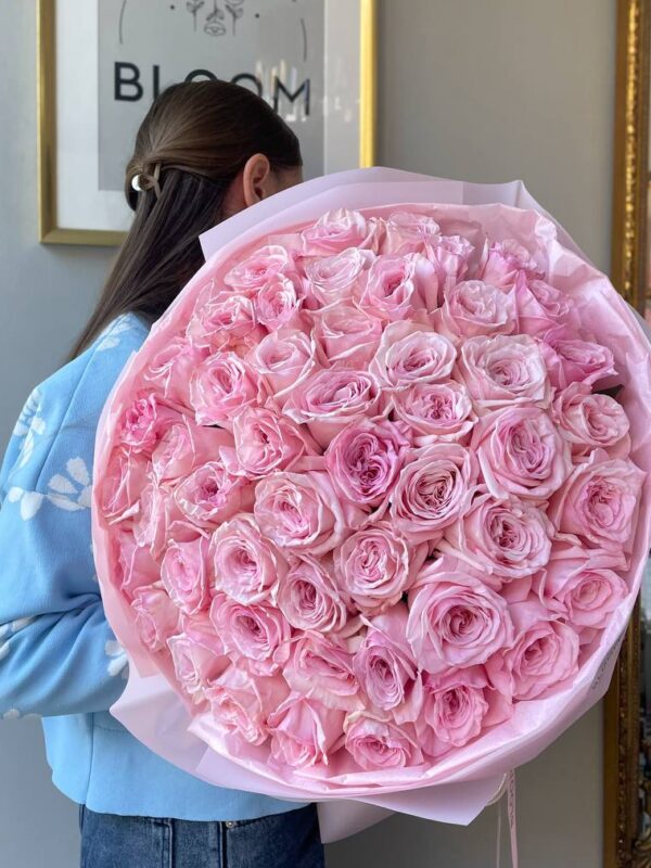 Великий моно-букет пудрово-рожевих троянд у рожевому пакуванні, який тримає дівчина, вид з-за спини.