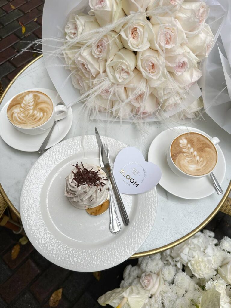 Преміальний букет кремово-рожевих троянд у фірмовій упаковці Bloom by Happy Doll на мармуровому столі поруч із десертом. Квіти Одеса доставка.