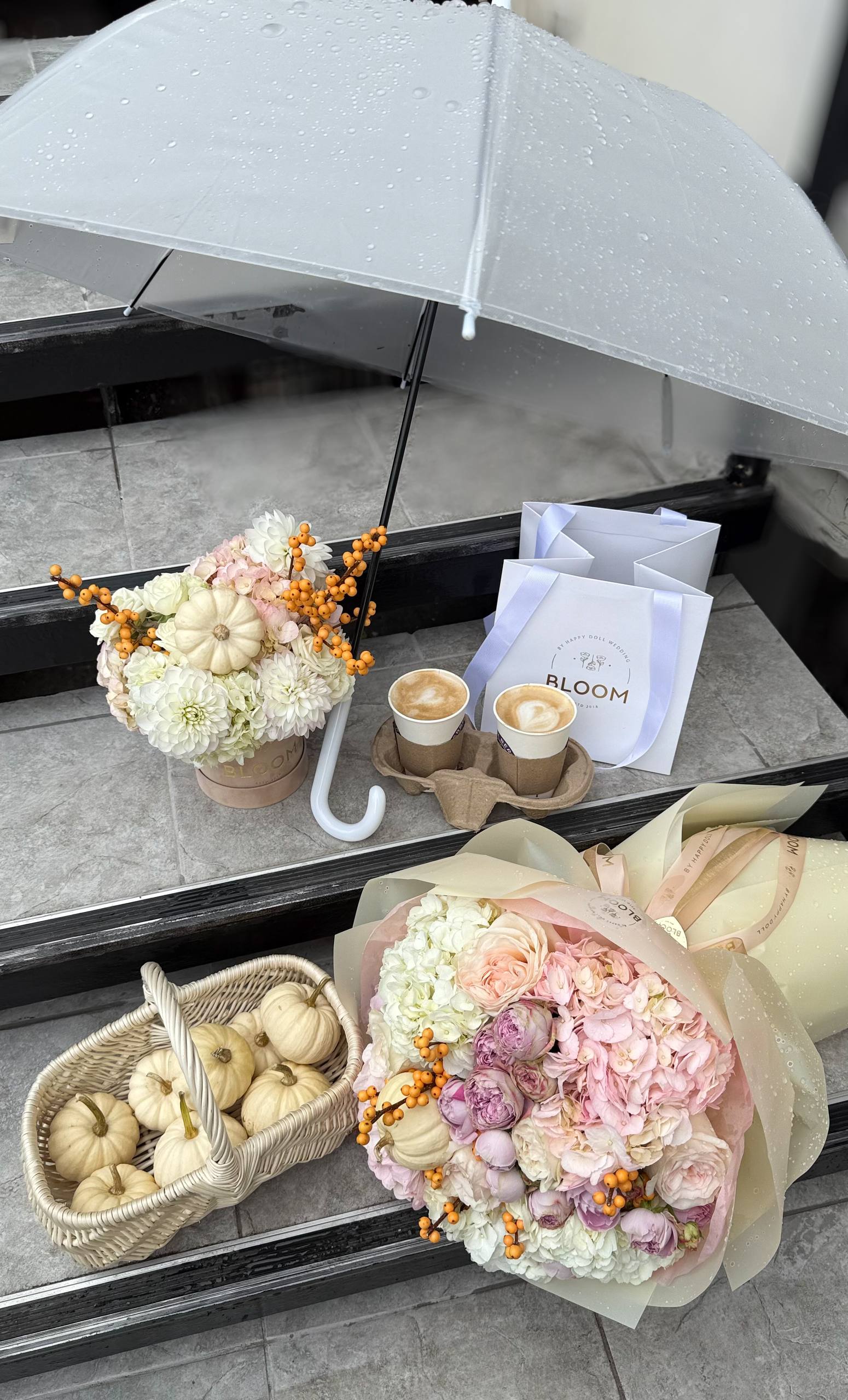 Осіння квіткова композиція Bloom by Happy Doll: авторські букети з гортензіями, півоніями, мінігарбузами та ягодами, розташовані поруч із кавою та прозорою парасолькою на дощових сходах.