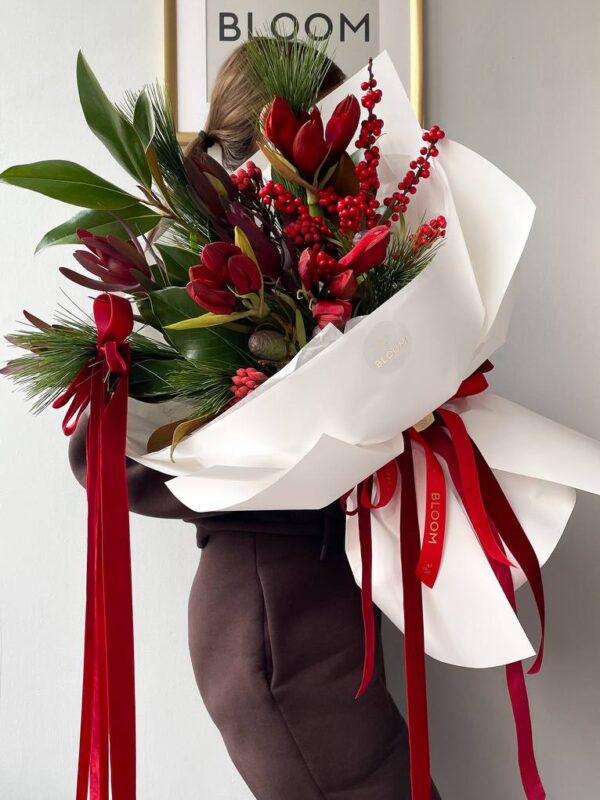 Зимовий святковий букет із червоними квітами, декоративними ягодами та ялиновими гілками, оформлений у білий папір із червоними стрічками.