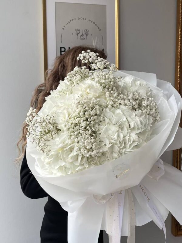 Букет із білої гортензії та гіпсофіли в ніжній упаковці поруч із елегантно загорнутими подарунками з білими та золотими стрічками.