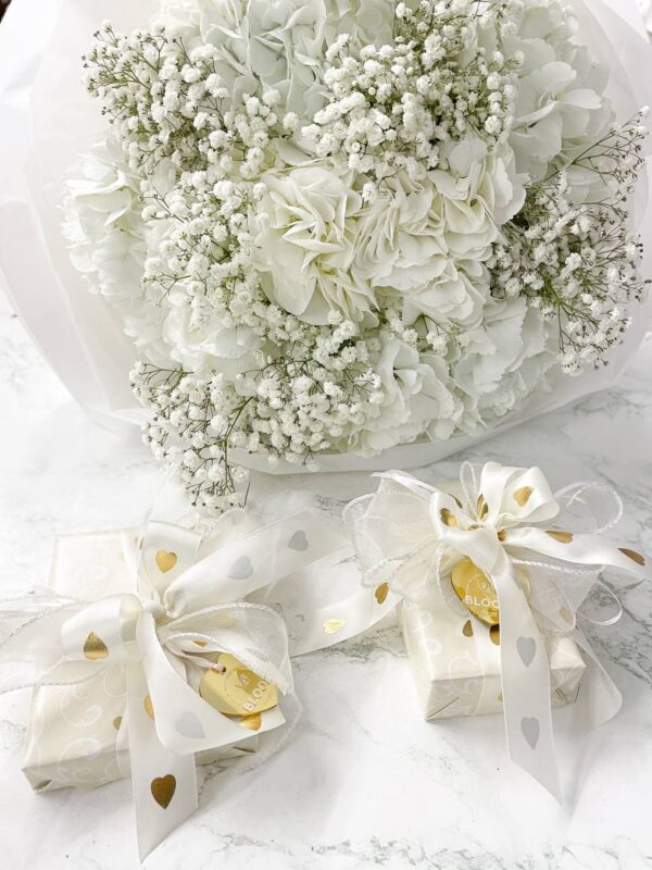 Frost and tenderness Букет із білої гортензії та гіпсофіли в ніжній упаковці поруч із елегантно загорнутими подарунками з білими та золотими стрічками.