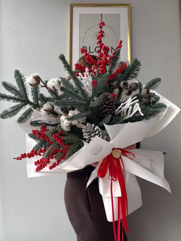 Зимова композиція з ялиновими гілками, білою бавовною, червоними ягодами та великими шишками, оформлена у білий папір із червоними стрічками.
