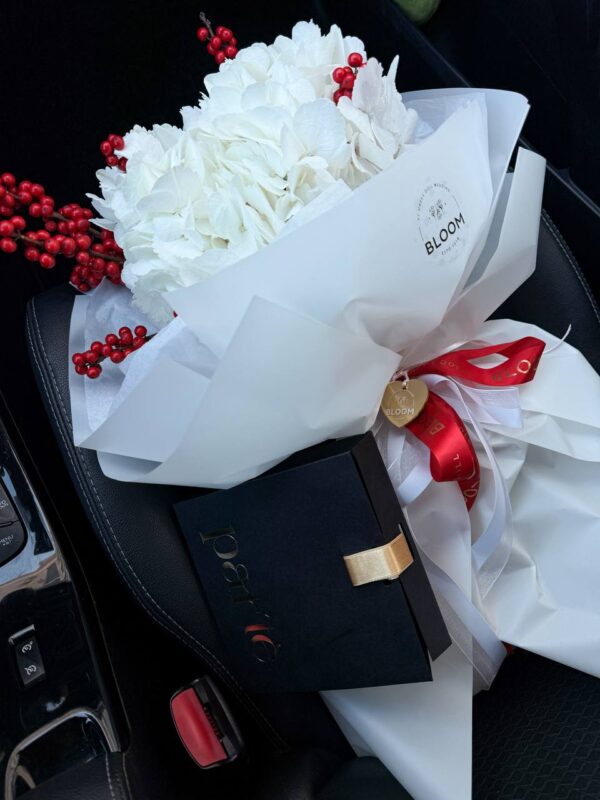 Білий букет з гортензії з червоними ягодами у фірмовому пакуванні Bloom, розміщений на сидінні автомобіля поруч із подарунковою коробкою.