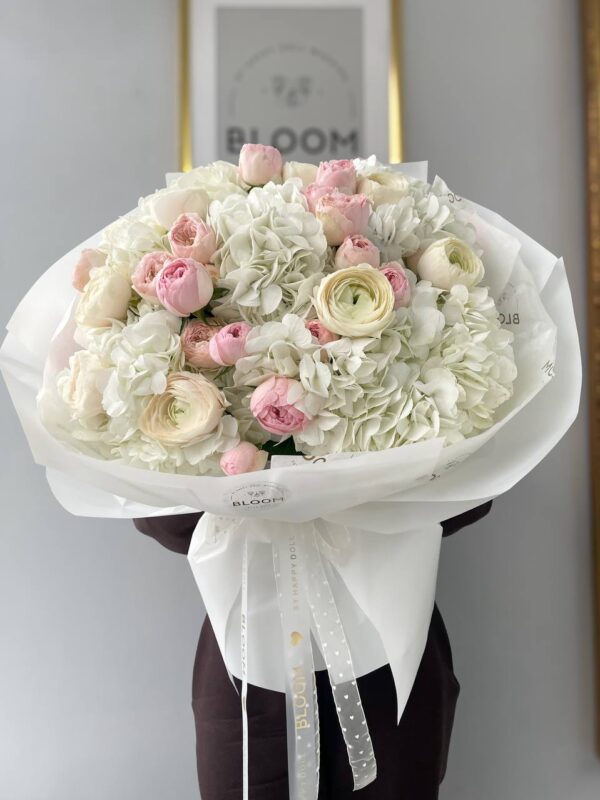 Ніжний букет квітів з білою гортензією, рожевими садовими трояндами та ранункулюсами у білій упаковці Bloom.