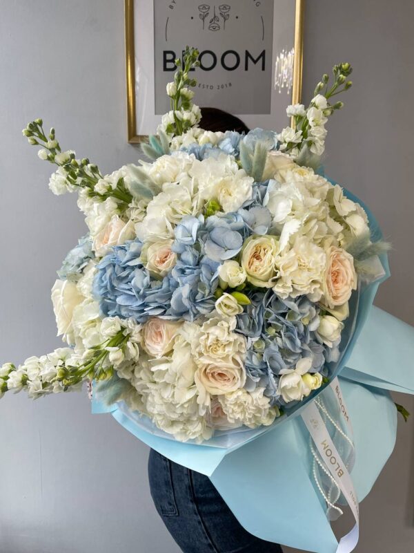 Букет з білої та блакитної гортензії, троянд O’Hara, білої фрезії та лагурусу в блакитному пакуванні Bloom.