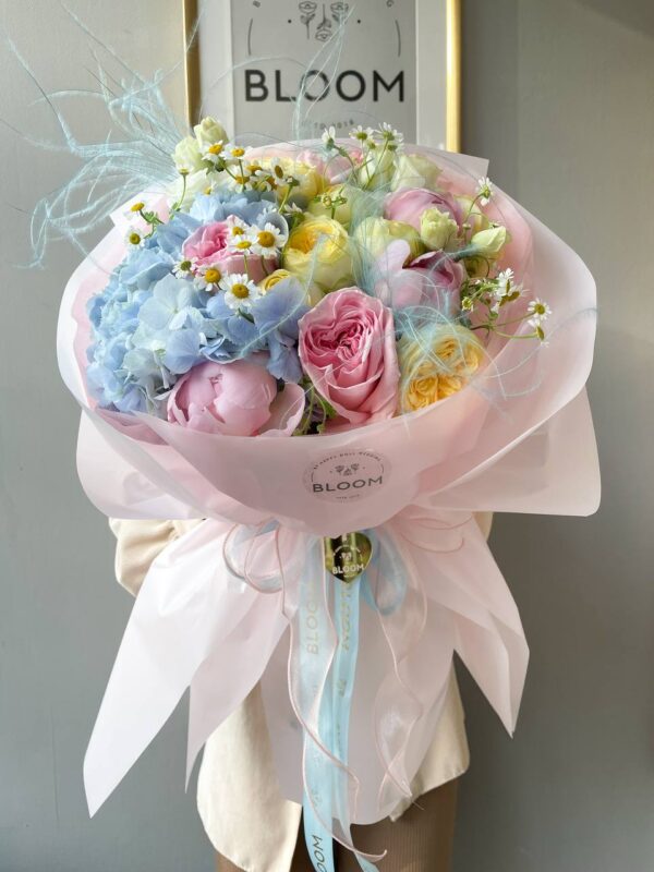 Букет у пастельних тонах з блакитною гортензією, рожевими трояндами, ранункулюсами та ромашками в упаковці Bloom.